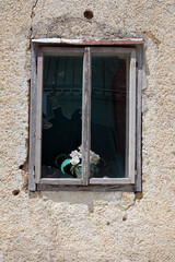 A weathered wooden window with cracked glass frames a faded bouquet of artificial white roses,...