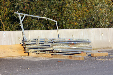 A neat stack of galvanized metal scaffolding pipes rests on wooden planks beside a concrete barrier...