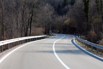 A smooth, curving asphalt road with white lane markings winds through a quiet, leafless forest,...