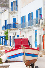 Red, white and blue fishing boat in front of white stone building with blue shutters on the island Levanzo in Italy.