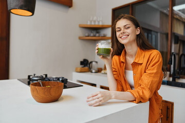 Happy young woman in an orange outfit enjoying a green smoothie in a modern kitchen with a wooden bowl, promoting healthy eating and wellness