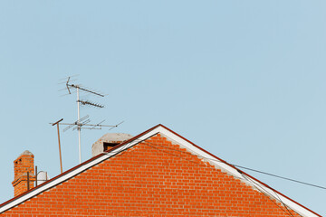 Minimalist architectural scene featuring the triangular red brick rooftop of a residential building with multiple old-style television antennas. A bright blue sky in the background emphasizes contrast