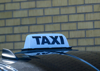 A taxi with a prominent TAXI sign is parked in front of a light brown brick wall. The setting appears to be a city area, likely during daytime, showcasing urban transport