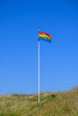 A vibrant rainbow flag flutters in the gentle breeze atop a tall pole. The clear blue sky contrasts beautifully with the colorful fabric. The surrounding grassy hillside adds to the serene atmosphere