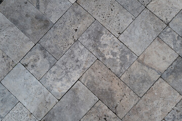 Close-up of a stone tile surface laid in a herringbone pattern, featuring varied textures, pores, and natural shades of gray and beige. A minimal and architectural composition rich in surface detail.