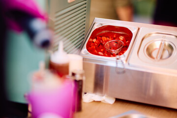 Close-up of a stainless steel soup warmer with a fruit-filled, red-colored beverage or sauce, accompanied by condiment bottles, set on a wooden table with a blurred background