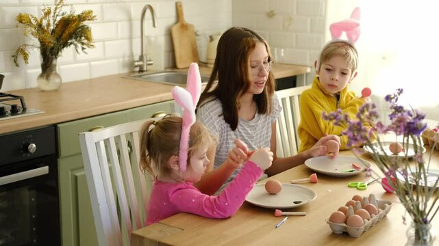Children siblings painting Easter egg sitting together at table. Easter Family traditions. Teen girl teaching happy little brother and sister to dye, decorate eggs with paints. Preparation to holidays