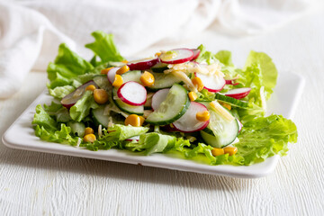 Vegan spring organic salad with fresh radish, cucumber, sweet corn and lettuce leaves with olive oil on white square plate. Healthy diet