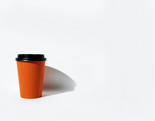 A simple image of an orange disposable coffee cup with a black lid placed on a white surface, casting a shadow to the left side, emphasizing minimalism and contrast