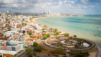 Drone photo of João Pessoa waterfront, Paraíba, Brazil, showing turquoise sea, boats, and urban beach skyline.