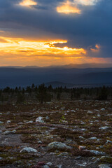 A breathtaking fiery sunset casts golden hues across the rugged mountains of Sweden and Norway. Heavy clouds loom above as the landscape darkens, revealing rocky terrain below