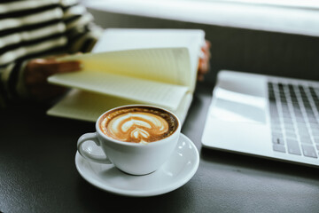 Young Asian woman making latte in a relaxed atmosphere with laptop and notebook and notepad while combining work and relaxation during a quiet coffee break at home or office.