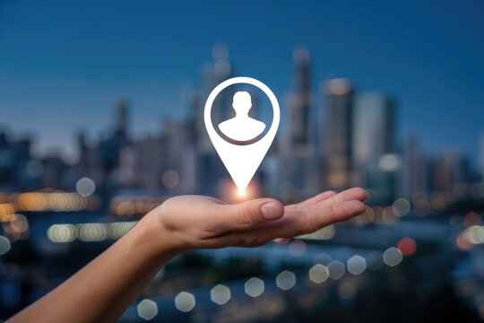 Hand holding a location pin with a person icon against a blurred city skyline at dusk