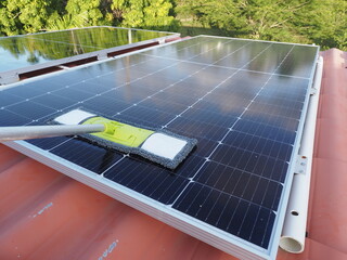 Worker uses a long-handled mop to clean solar panels installed on a rooftop. This maintenance helps...