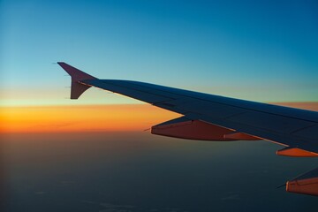 Fototapeta premium An airplane wing slices through a vibrant sunset. The sky shifts from deep blue to fiery orange, creating a stunning aerial view, perfect for themes of travel and adventure.