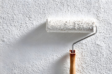 Close-up of a paint roller applying white paint to a textured wall, showing details of the roller, wall, and paint application in a studio shot.