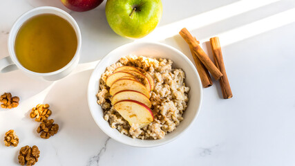 A nutritious bowl of oatmeal topped with sliced apples and cinnamon, served with a cup of tea, walnuts, and whole apples on a marble surface.