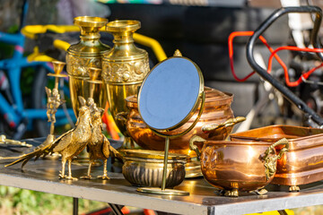 Antique market stall with golden and copper vessels, decorative bird figurines, and a round mirror on a sunny day.