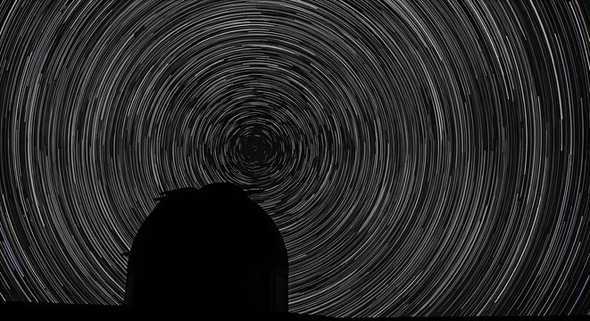 Ethereal star trails illuminate the night sky above a silhouette of the observatory building, creating a cosmic backdrop.