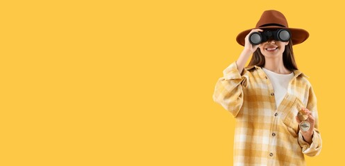 Female tourist with compass looking through binoculars on yellow background with space for text © Pixel-Shot