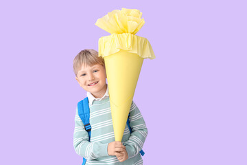 Happy little boy with backpack and yellow school cone on lilac background