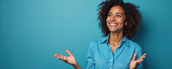Confident Brazilian woman smiles broadly, presenting idea with open hands against solid blue background. Curly hair frames face as gestures to empty space, suggesting communication, positive