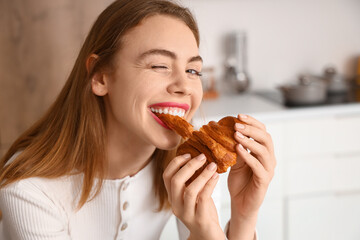 Beautiful young woman eating tasty croissant in kitchen