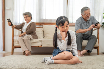 Asian girl sad sitting alone on floor in living room. Grandfather and grandmother addicted to smartphones and do not care about their granddaughter. Problems in family and bad relations.