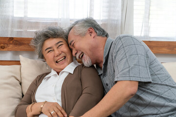 Asian elderly couple enjoying joyful moment together at home, smiling and laughing while sitting closely on a cozy sofa