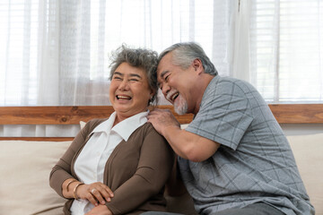 Asian elderly couple enjoying joyful moment together at home, smiling and laughing while sitting closely on a cozy sofa