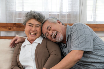 Asian elderly couple enjoying joyful moment together at home, smiling and laughing while sitting closely on a cozy sofa