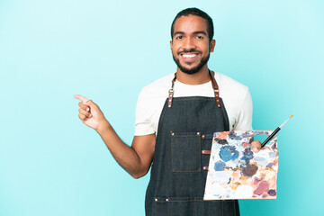 Young artist latin man holding a palette isolated on blue background pointing finger to the side