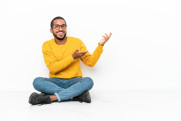 Young Ecuadorian man sitting on the floor isolated on white wall extending hands to the side for...