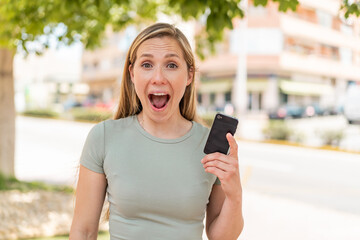 Young blonde woman using mobile phone at outdoors with surprise and shocked facial expression