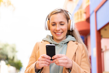 Young blonde woman at outdoors listening music and looking to mobile