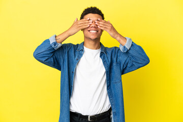 Young African American man isolated on yellow background covering eyes by hands and smiling