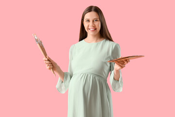 Young pregnant female artist with brushes and palette on pink background