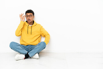 Young African American man sitting on the floor isolated on white background with glasses and surprised