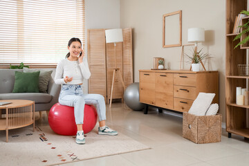 Young Asian woman talking by mobile phone on fitball at home