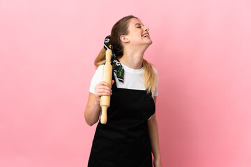 Young blonde woman holding a rolling pin isolated on pink background laughing in lateral position