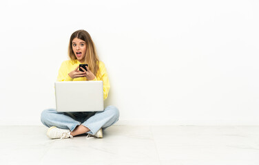 Young woman with a laptop sitting on the floor surprised and sending a message
