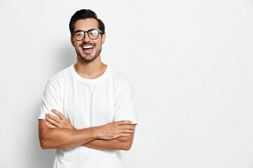 Happy smiling young man in casual white t-shirt wearing glasses, crossed arms, looking at camera...