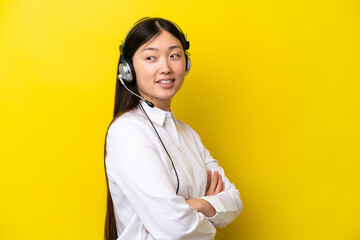 Telemarketer Chinese woman working with a headset isolated on yellow background with arms crossed...