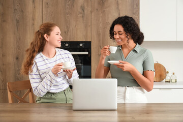 Beautiful young happy female friends with cups of coffee and laptop sitting in kitchen at home