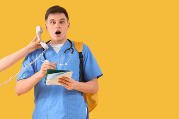 Shocked medical intern with notebook receiving phone on yellow background
