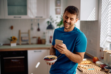 Happy man using cell phone while having breakfast in kitchen.