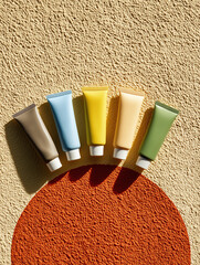 Assorted cosmetic tubes in pastel tones lined up on textured surface with warm daylight and bold orange graphic circle shadow design.