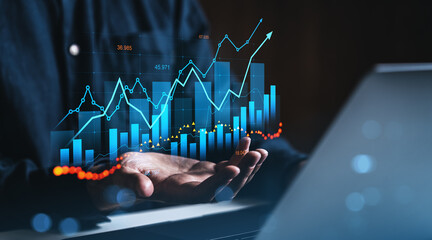 Businessman holding holographic financial chart with upward growth lines and bar graphs above hands in dark office environment, symbolizing analytics concept.