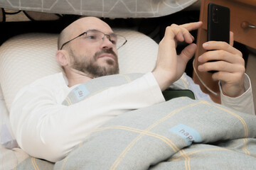 Man relaxing comfortably in bed using his smartphone. Casual indoor lifestyle scene showing technology, digital connection, and leisure time at home.