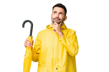 Young caucasian man with rainproof coat and umbrella over isolated background having doubts and...